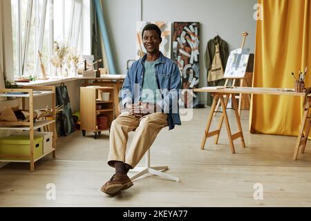 Young professional painter or teacher of arts sitting in spacious studio against his workplace with easel, table and canvases Stock Photo