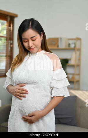 Portrait, Beautiful and happy young Asian pregnant woman stands in living room, touching her belly with love, feeling her baby in her stomach. motherh Stock Photo