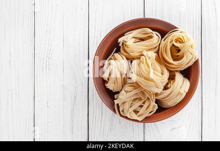 Uncooked tagliatelle pasta in ceramic bowl on white wooden background with copy space Stock Photo