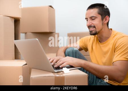 Happy man in his empty new home apartment among the boxes with laptop moving house online trading Stock Photo