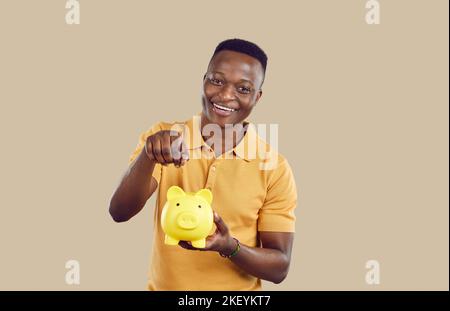Cheerful young African American man tossing money into piggy bank making savings stands in studio Stock Photo
