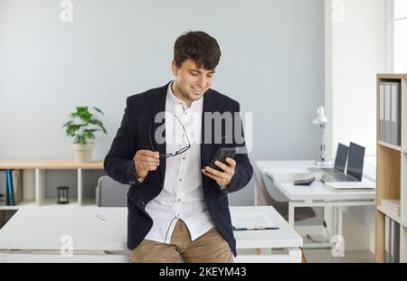 Young businessman standing in office and using online banking app on mobile phone Stock Photo