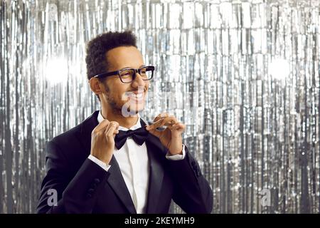 Happy handsome young ethnic man in tuxedo and stylish glasses adjusting his bow tie Stock Photo