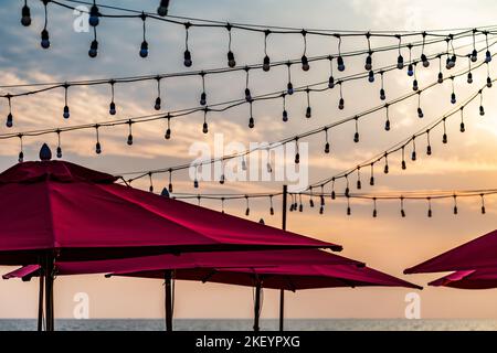 The top part of many beach red umbrellas and many party light bulbs hang in the air, colorful orange sky over the sea. Stock Photo