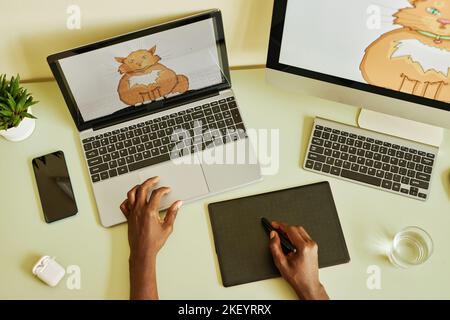 Above angle of hands of young black man holding stylus over graphic tablet while drawing fat fluffy ginger cat by workplace in office Stock Photo