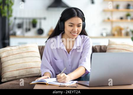female student studying remotely using a laptop, taking notes on notepad during online lesson, e-learning Stock Photo