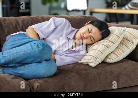 woman suffering from abdominal pain lying on the couch at home Female suffering from severe spasms holding her stomach Stock Photo