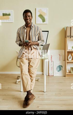 Happy young creative freelancer in casualwear using smartphone while standing against workplace with desktop computers in office Stock Photo