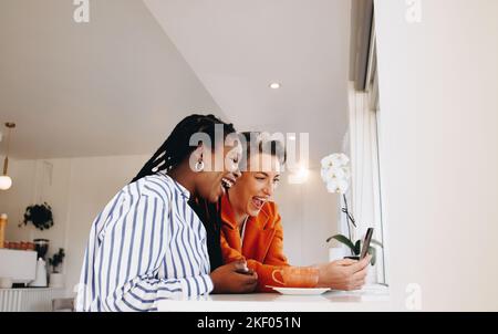 Two happy business women laughing excitedly while using a smartphone together in a cafe. Cheerful female colleagues enjoying their coffee break togeth Stock Photo