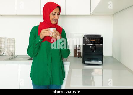Happy mature islamic woman enjoying fresh aromatic coffee near modern machine in kitchen interior Stock Photo