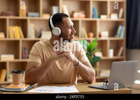 Positive pensive arab man working online at home, sitting at workdesk in front of laptop, looking aside at copy space Stock Photo