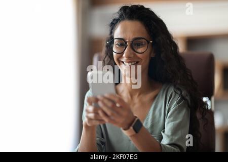 Cheerful brunette woman sitting at workdesk, using smartphone Stock Photo