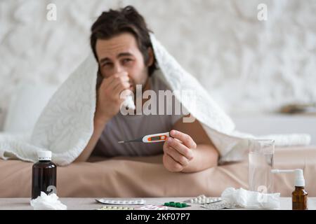 Unhappy millennial european man with stubble blows nose in napkin, lies on bed under blanket, show thermometer Stock Photo