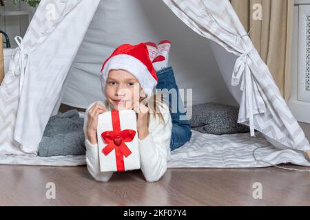 A girl in a Santa hat holds his big gift box for Christmas New Year lying in a children's tent wigwam in the nursery. Christmas gifts for children. Fa