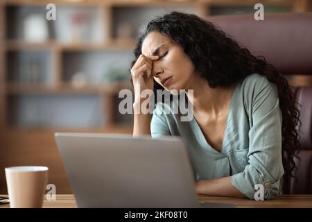 Exhausted young female employee working on laptop, touching nose bridge Stock Photo