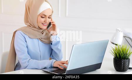 Muslim woman in hijab is talking mobile phone and typing on laptop at office work. Stock Photo