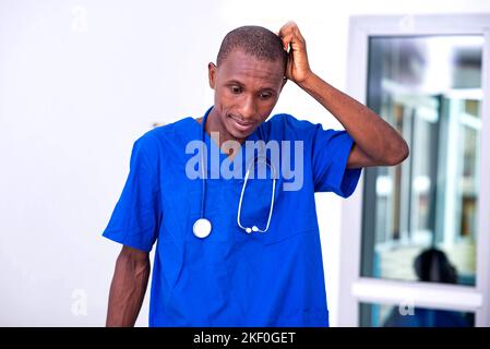 portrait of confused young male doctor, wearing blue uniform at hospital, scratching his head. Stock Photo