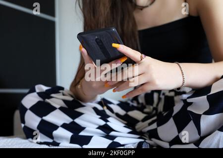 Smartphone in hands of girl with orange acrylic nails sitting on a sofa. Female manicure, online communication, using mobile phone in home Stock Photo