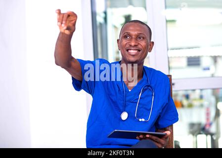 young male doctor sitting on a chair, holding a digital tablet and gesturing his hand while smiling. Stock Photo