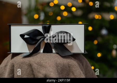 Christmas gift with the black ribbon on the table. Blurred lights in the background, white present box with dark strip. Stock Photo