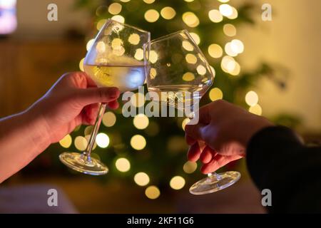Hands holding glass of wine, blurry christmas lights on the christmas tree in the background. Pine tree decorated with shining garland, two persons ho Stock Photo