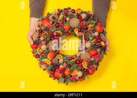 Hands holding a colorful wreath in the yellow background. Garland decorated with fruit and berries on the bright surface in persons hands. Stock Photo