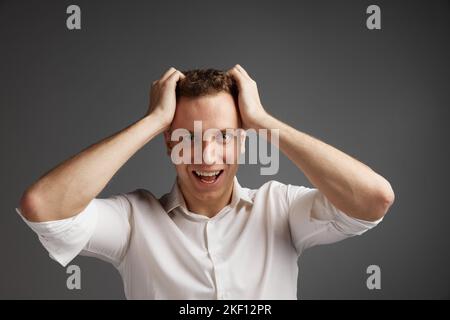 Portrait of young man emotionally posing in white shirt isolate over grey studio background. Shocked, surprise Stock Photo