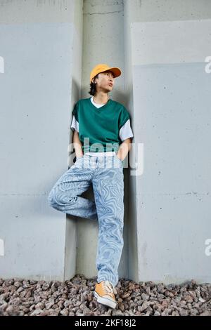 Vertical portrait of young Asian man wearing colorful street style clothes standing by concrete wall in urban setting Stock Photo