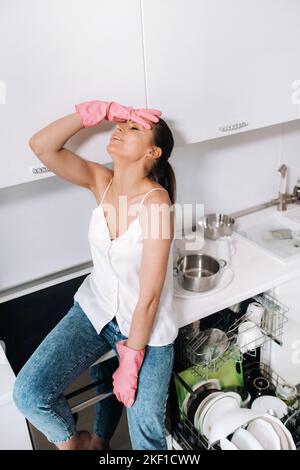 a housewife girl in pink gloves after cleaning the house sits tired in the kitchen.In the white kitchen, the girl has washed the dishes and is resting Stock Photo