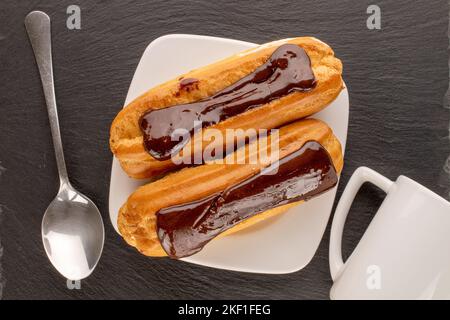 Two sweet eclairs with white plate, spoon and cup on slate stone, macro, top view. Stock Photo