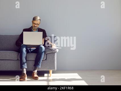 Mature businessman working on laptop while sitting on sofa near grey copy space wall Stock Photo