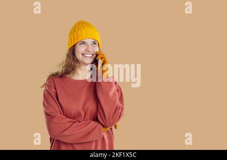Smiling cheerful young woman in yellow hat and gloves is calling on smartphone on beige background. Stock Photo