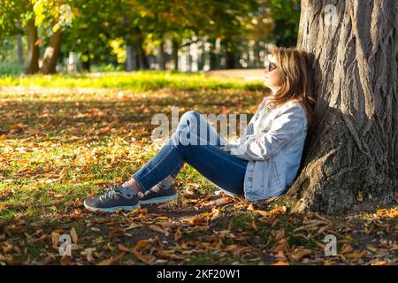 Mature white woman sitting on the grass of a park and leaning on a tree trunk in a relaxed way. Stock Photo
