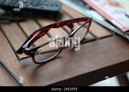 A portrait of a pair of brown reading glasses lying on a wooden table next to some magazines. The specs are ready to be used to read or watching telev Stock Photo