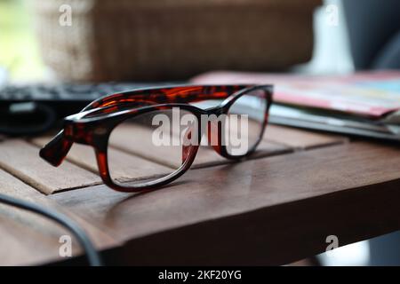 A close up portrait of a pair of brown reading glasses lying on a wooden table next to some magazines. The specs are ready to be used to read or watch Stock Photo