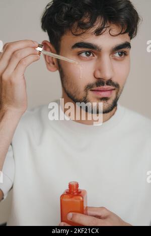 Close-up image of handsome man holding pipette with oil, applying serum beauty product on his face. Isolated over gray background. Organic oil. Body Stock Photo