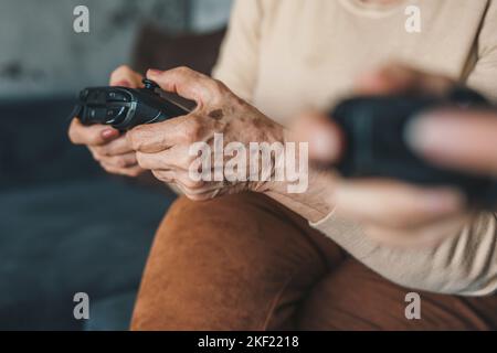 Senior woman's hands holding joysticks sitting cozy on the sofa in bright sunny living room. Senior playing console for lifestyle design. Stock Photo