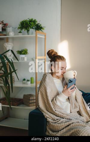 Caucasian woman wrapped in warm cozy plaid holding mug of tea, enjoying leisure at home while chatting with someone on mobile phone. Domestic Stock Photo