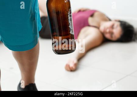 man with a beer bottle in his hand, leaving his wife lying on the floor after having beaten her. domestic violence and abuse of women Stock Photo