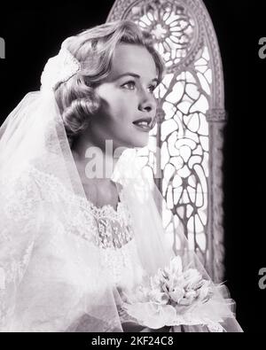 1960s YOUNG WOMAN BRIDE WEARING VEIL HOLDING BOUQUET IN CHURCH IN FRONT OF STAINED GLASS WINDOW - b16035 HAR001 HARS MARRIAGE PERSONS VEIL CONFIDENCE B&W BRIDAL DREAMS HAPPINESS BRIDES EXCITEMENT LOW ANGLE NUPTIAL NUPTIALS MARRYING PRIDE IN OF CONCEPTUAL RITE OF PASSAGE STYLISH WED MARRY MATRIMONY YOUNG ADULT WOMAN BLACK AND WHITE CAUCASIAN ETHNICITY HAR001 OLD FASHIONED Stock Photo