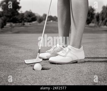 1960s CLOSE-UP OF FEMALE LEGS WEARING WHITE GOLF SHOES AND CLUB PUTTER ABOUT TO STROKE BALL INTO HOLE CUP ON THE GREEN - g6856 HAR001 HARS SINKING WOMAN'S GOALS SUCCESS COURSE GOLFERS LEISURE LOW ANGLE RECREATION INTO CONCEPTUAL ATHLETES CLOSE-UP LINKS RECREATION GOLFING TAPPING GROWTH MID-ADULT MID-ADULT WOMAN PRECISION PUTT PUTTER BLACK AND WHITE CAUCASIAN ETHNICITY HAR001 KNEE DOWN LEGS ONLY OLD FASHIONED PAR PUTTING GREEN STROKING THE GREEN Stock Photo