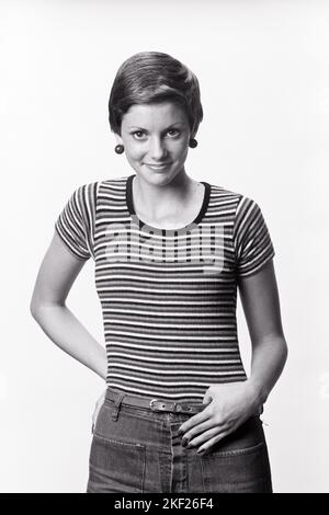 1970s TRENDY YOUNG WOMAN SHORT HAIRCUT WEARING ROUND BAUBLE EARRINGS HIP-HUGGER JEANS STRIPED T-SHIRT SMILING LOOKING AT CAMERA  - g8375 HAR001 HARS FEMALES STRIPED STUDIO SHOT MODERN HAIRCUT TEE COPY SPACE HALF-LENGTH LADIES PERSONS EARRINGS STRIPES TRENDY CONFIDENCE DENIM B&W EYE CONTACT HAPPINESS CHEERFUL STYLES SMILES JOYFUL STYLISH TEENAGED T-SHIRT BLUE JEANS FASHIONS INFORMAL TWILL YOUNG ADULT WOMAN BLACK AND WHITE CASUAL CAUCASIAN ETHNICITY HAR001 OLD FASHIONED Stock Photo