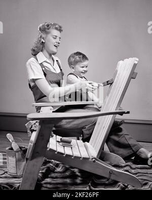 1940s 1950s SMILING WOMAN MOTHER PAINTING ADIRONDACK CHAIR WITH HELP FROM BOY HER LAUGHING TODDLER SON  - j10020 HAR001 HARS PAIR DOMESTIC HER MOTHERS OLD TIME NOSTALGIA OLD FASHION 1 HOUSEHOLD SILLY JUVENILE STYLE COMMUNICATION YOUNG ADULT COMIC TEAMWORK STRONG SONS PLEASED JOY LIFESTYLE SATISFACTION CHORES FEMALES STUDIO SHOT HOME LIFE COPY SPACE FRIENDSHIP HALF-LENGTH LADIES PERSONS CARING MALES ADIRONDACK B&W GOALS HUMOROUS HAPPINESS CHEERFUL CHORE COMICAL PRIDE SMILES TASKS UPDO CONNECTION VICTORY ROLLS COMEDY JOYFUL BABY BOY DIY PERSONAL ATTACHMENT AFFECTION COOPERATION CREATIVITY Stock Photo