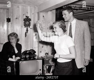 1950s TEEN BOY GIVING TEENAGE GIRL HANDS ON HELP AIMING A DART DURING PARTY IN KNOTTY PINE REC ROOM ANOTHER GIRL LEANING ON TV - j7020 HAR001 HARS OLD TIME NOSTALGIA OLD FASHION 1 GIVING JUVENILE STYLE COMMUNICATION TEAMWORK PINE PLEASED JOY LIFESTYLE SATISFACTION FEMALES HEALTHINESS HOME LIFE COPY SPACE FRIENDSHIP HALF-LENGTH PERSONS MALES TEENAGE GIRL TEENAGE BOY CONFIDENCE B&W DATING GOALS SUIT AND TIE HAPPINESS CHEERFUL LEISURE EXCITEMENT REC RECREATION ATTRACTION PARTIES RELATIONSHIPS SMILES CONNECTION COURTSHIP DART JOYFUL STYLISH SUPPORT TEENAGED LEANS POSSIBILITY AIMING ANOTHER Stock Photo