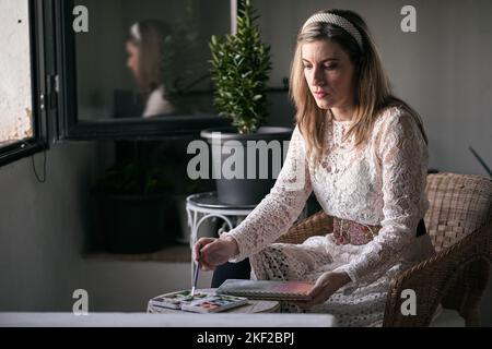 blond caucasian mature woman sitting in a wicker armchair painting with watercolors and a fine brush in a notebook by the window Stock Photo