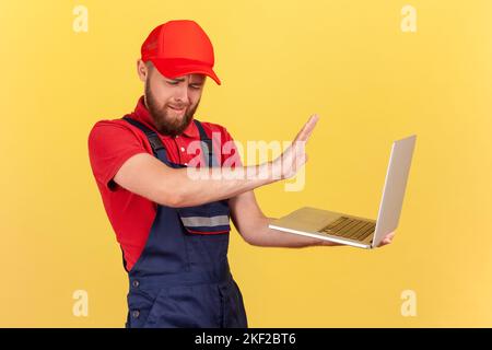 Side view portrait of handsome worker man wearing blue uniform and red cap holding notebook, forbidden information, showing stop ban gesture. Indoor studio shot isolated on yellow background. Stock Photo