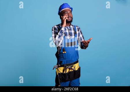 African american construction worker having a serious mobile phone conversation with materials supplier. Builder talking on smart phone wearing coverall and hard hat, studio shot. Stock Photo