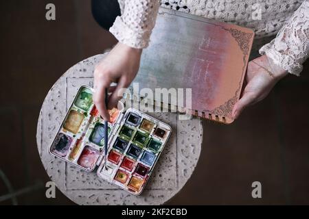 closeup of a case of watercolors of many colors and a hand holding a fine brush ready to paint on a notebook Stock Photo