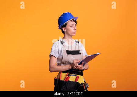 Construction worker taking notes and measurements to work on renovating project, holding clipboard papers in studio. Young supervisor engineer analyzing information on files, industrial skills. Stock Photo
