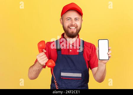 Portrait of delighted smiling worker man holding out retro phone and cell phone with empty screen for advertisement, choose best device for you, Indoor studio shot isolated on yellow background. Stock Photo